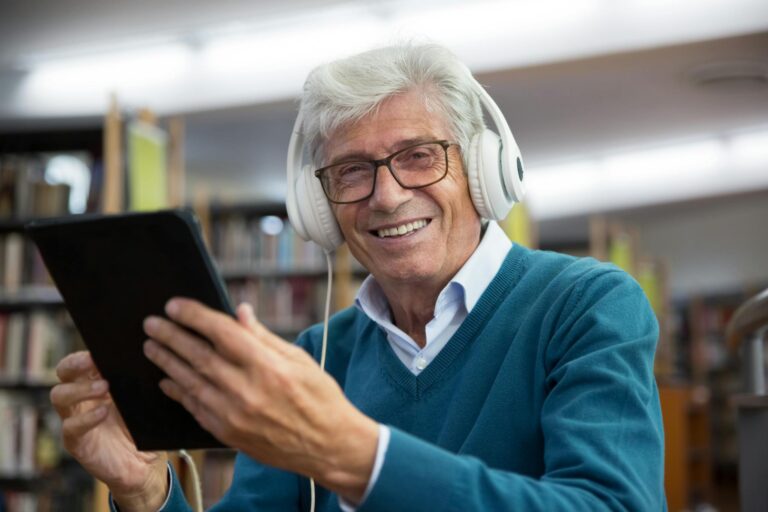 Senior man wearing headphones using a tablet in a library. Happy expression showcases enjoyment of technology.