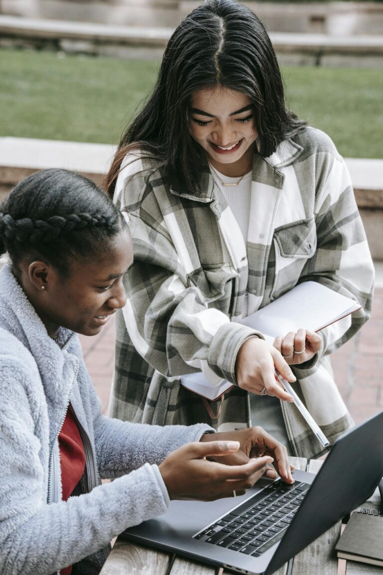 Young smiling Asian woman showing at netbook near black girlfriend while studying at table in town